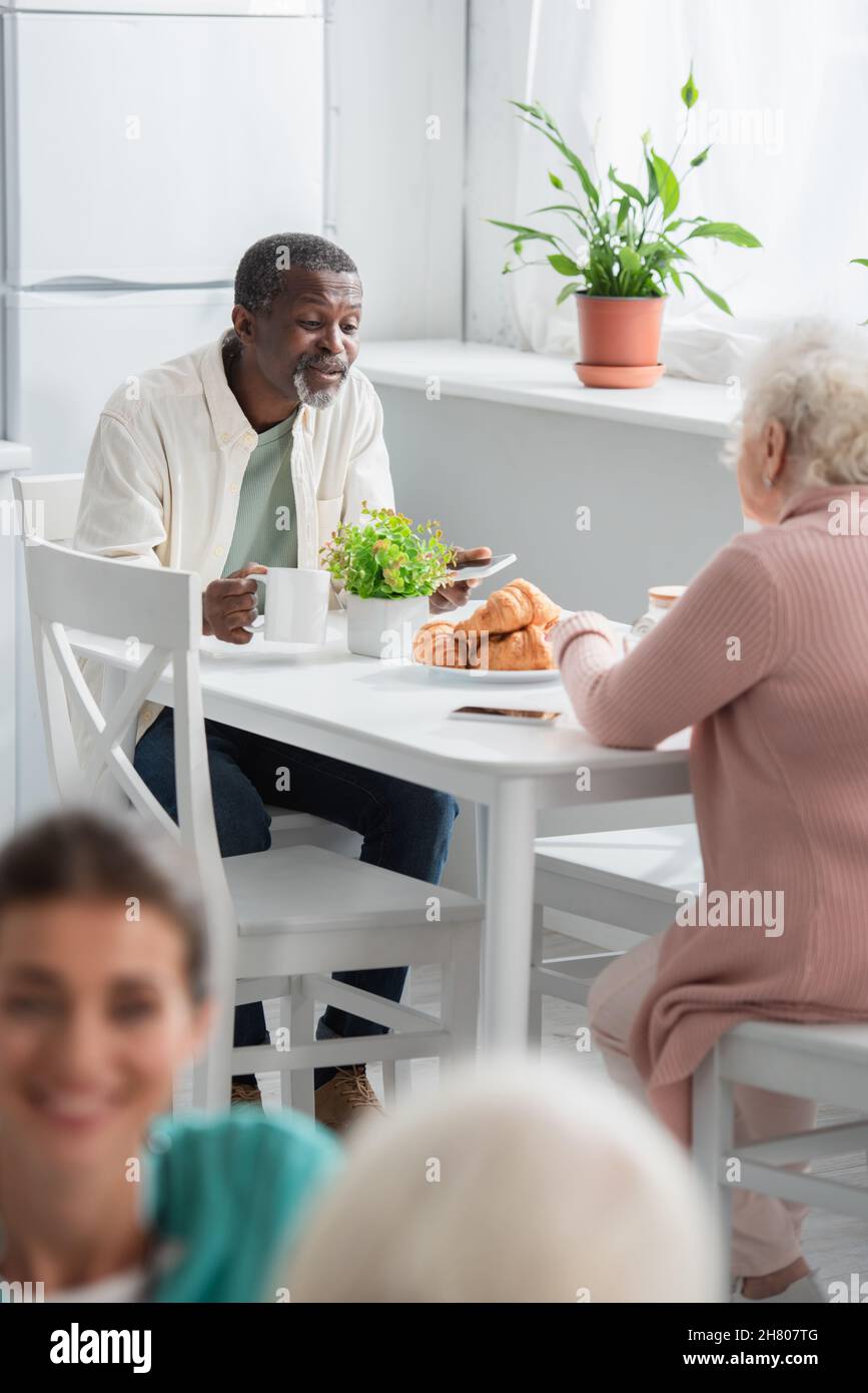 Uomo afro-americano con tè e smartphone parlare con un amico in casa di cura Foto Stock