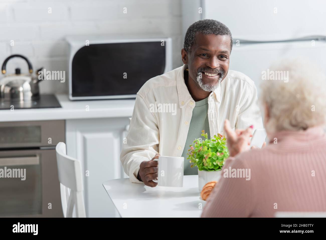 Uomo afro-americano positivo che tiene il tè vicino a un amico sfocato in casa di cura Foto Stock