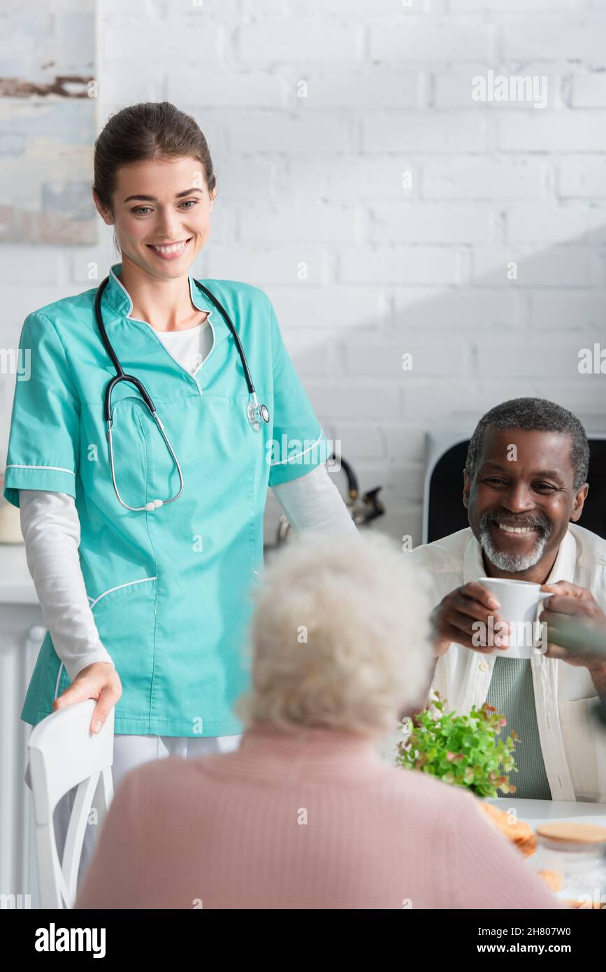 Infermiera sorridente in piedi vicino a pazienti interrazziali in casa di cura Foto Stock