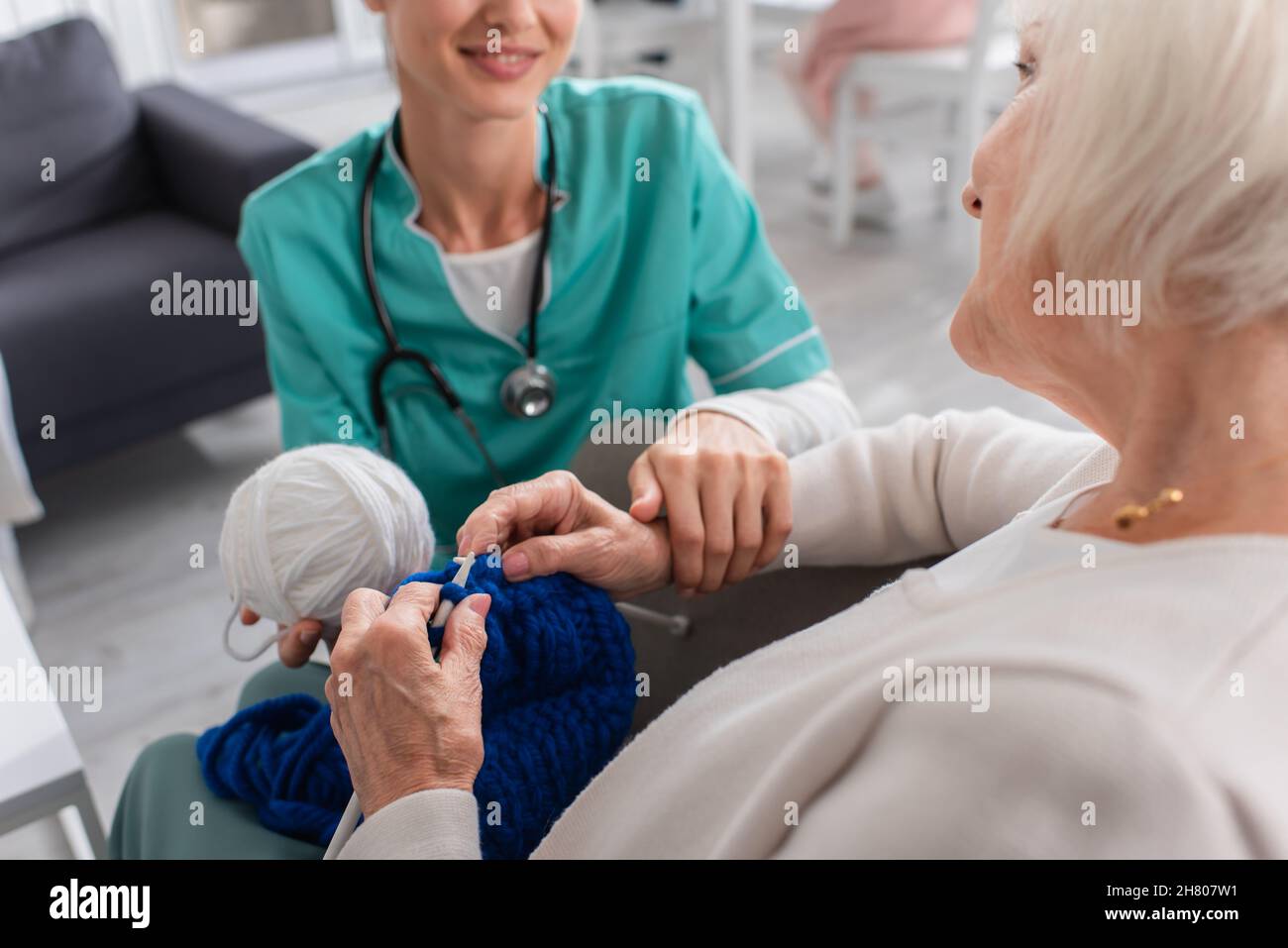 Donna anziana che si inginocchiava vicino a un'infermiera sorridente sfocata in uniforme in casa di cura Foto Stock