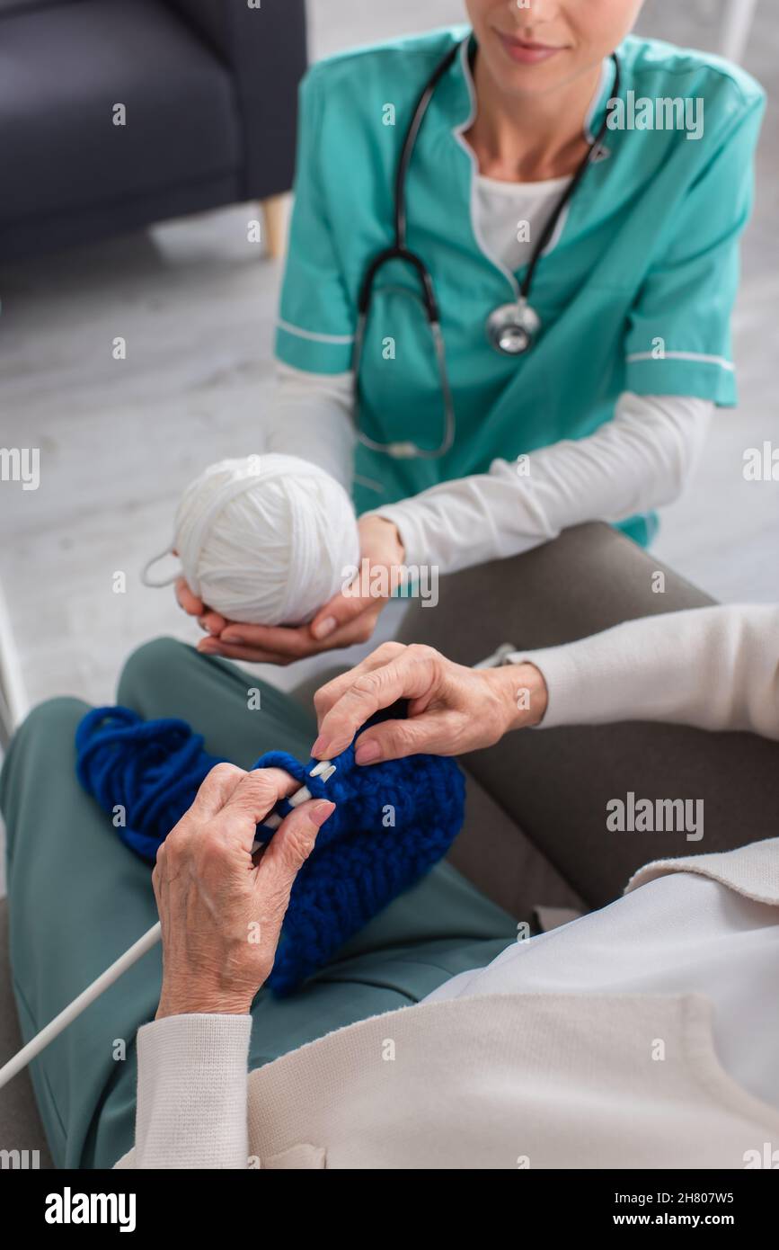 Vista croppata della donna anziana che si maglia vicino infermiere che tiene il filo in casa di cura Foto Stock