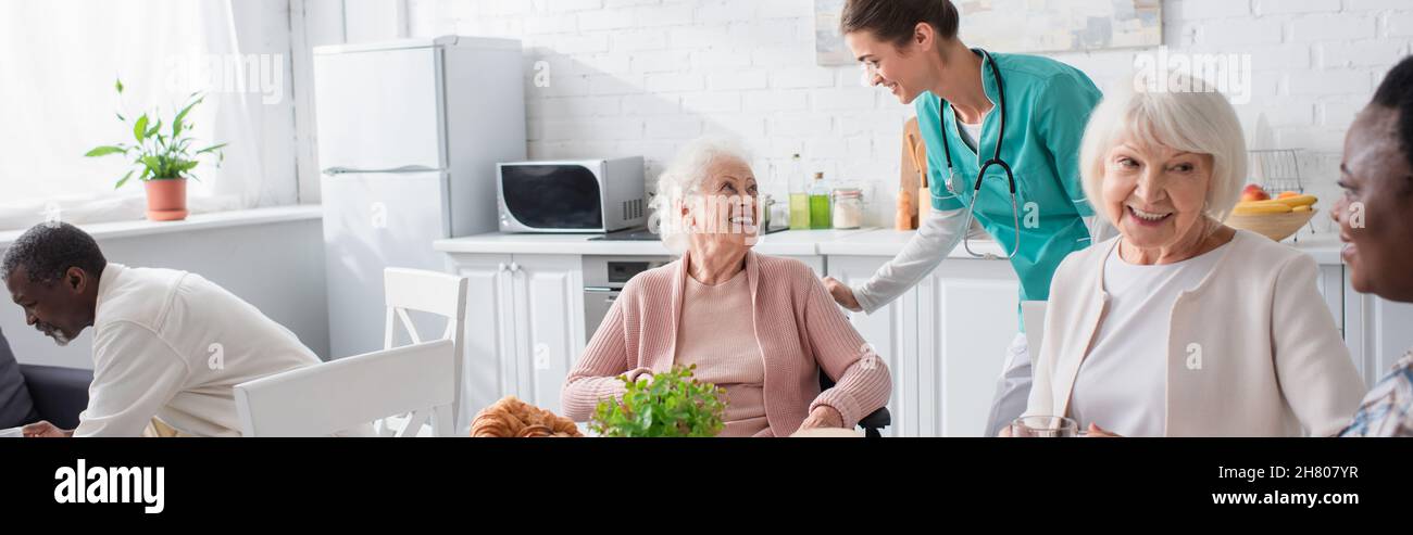 Infermiera che parla con le donne anziane in sedia a rotelle vicino ai pensionati interrazziali in casa di cura, banner Foto Stock