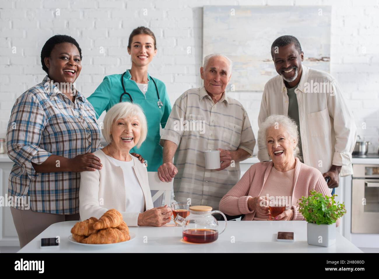 Infermiera sorridente in piedi vicino a pazienti interrazziali e tè in casa di cura Foto Stock