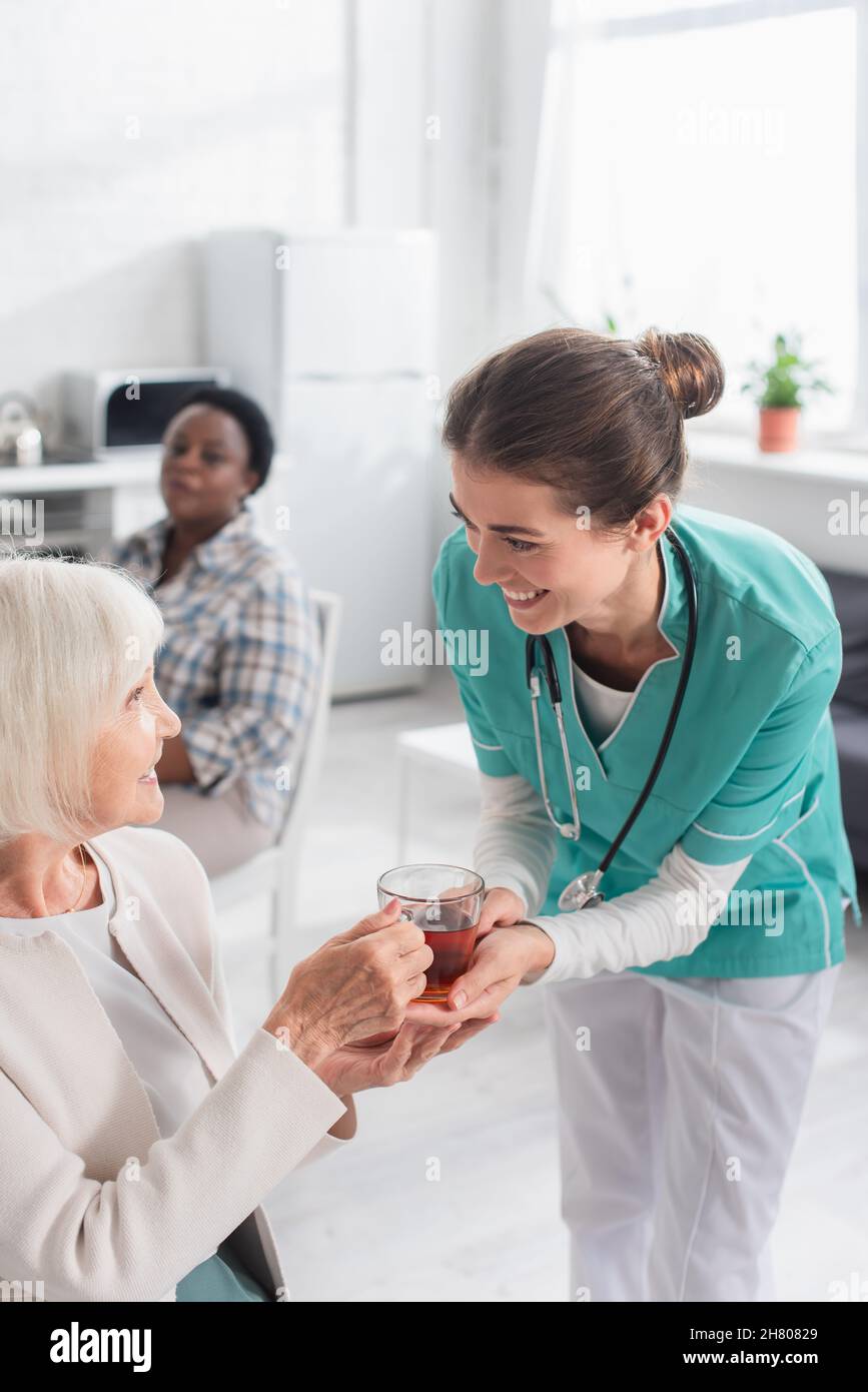 Infermiera sorridente che tiene il tè vicino al paziente nella casa di cura Foto Stock