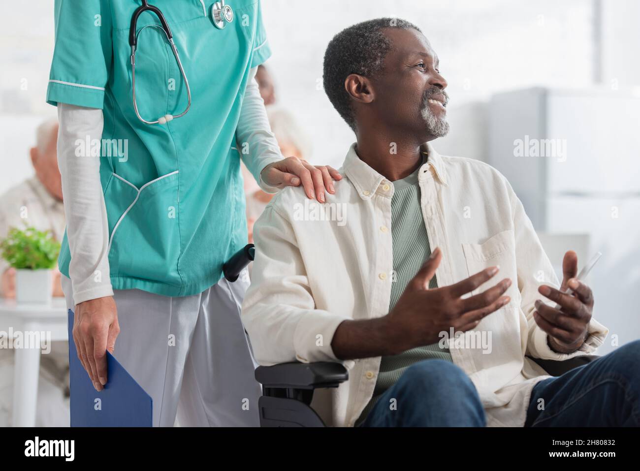 Sorridente paziente afroamericano che tiene lo smartphone mentre si siede in sedia a rotelle vicino all'infermiere in casa di cura Foto Stock