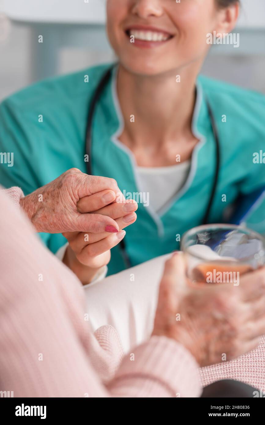 Vista ritagliata dell'infermiera sorridente che tiene la mano del paziente anziano con il tè nella casa di cura Foto Stock