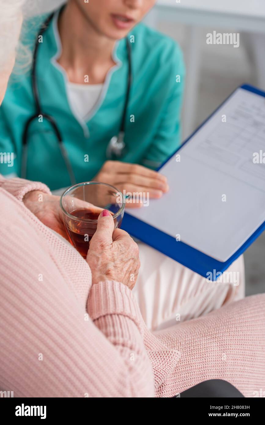 Vista croppata della donna anziana che tiene il tè vicino all'infermiera sfocata con gli appunti nella casa di cura Foto Stock
