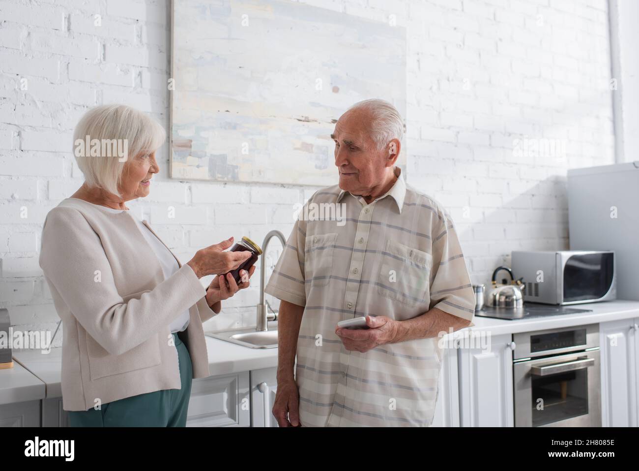 Donna anziana sorridente che punta al vaso con marmellata vicino ad un amico con smartphone in casa di cura Foto Stock