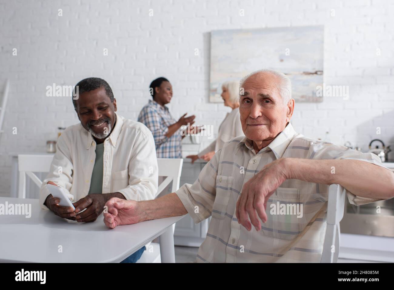 Uomo anziano che guarda la macchina fotografica vicino sorridente amico afroamericano con smartphone in casa di cura Foto Stock
