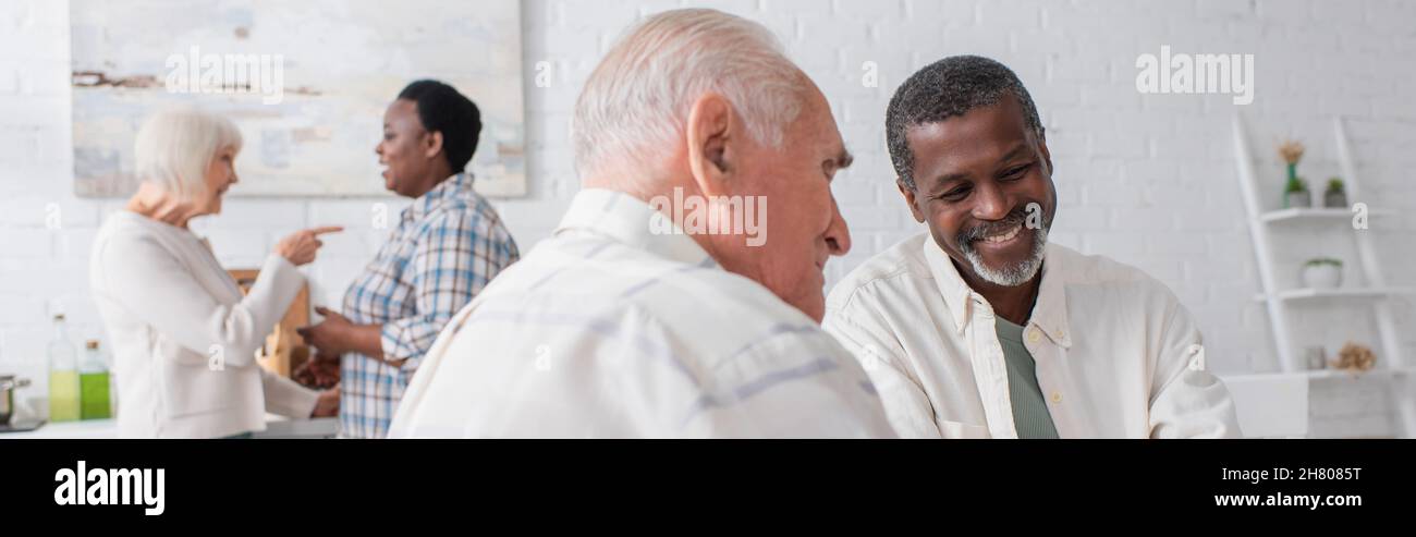 Sorridendo anziani uomini interrazziali parlare vicino donne sfocate in casa di cura, banner Foto Stock