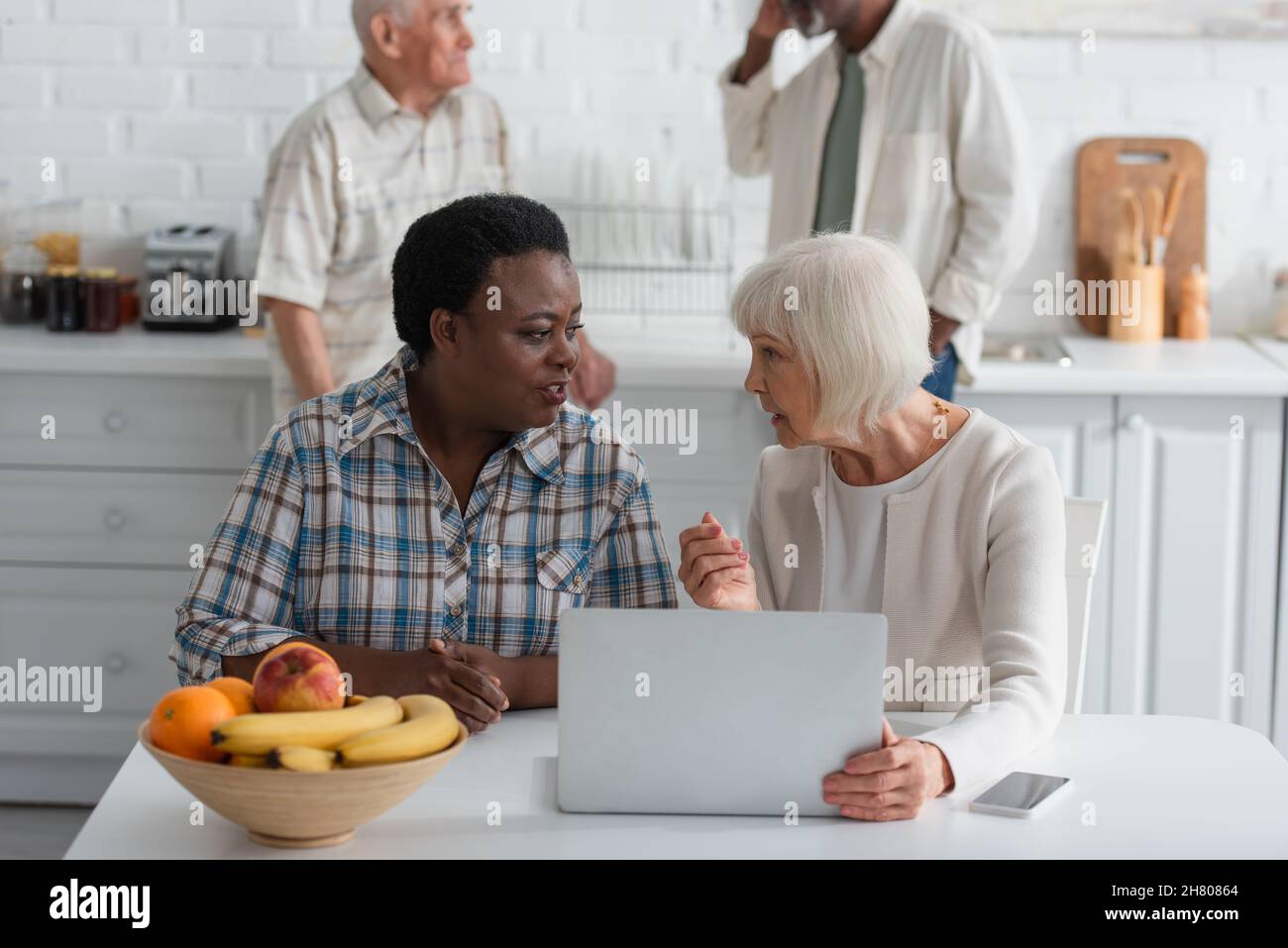 Donne interracial anziane che parlano vicino ai dispositivi e ai frutti nella casa di cura Foto Stock