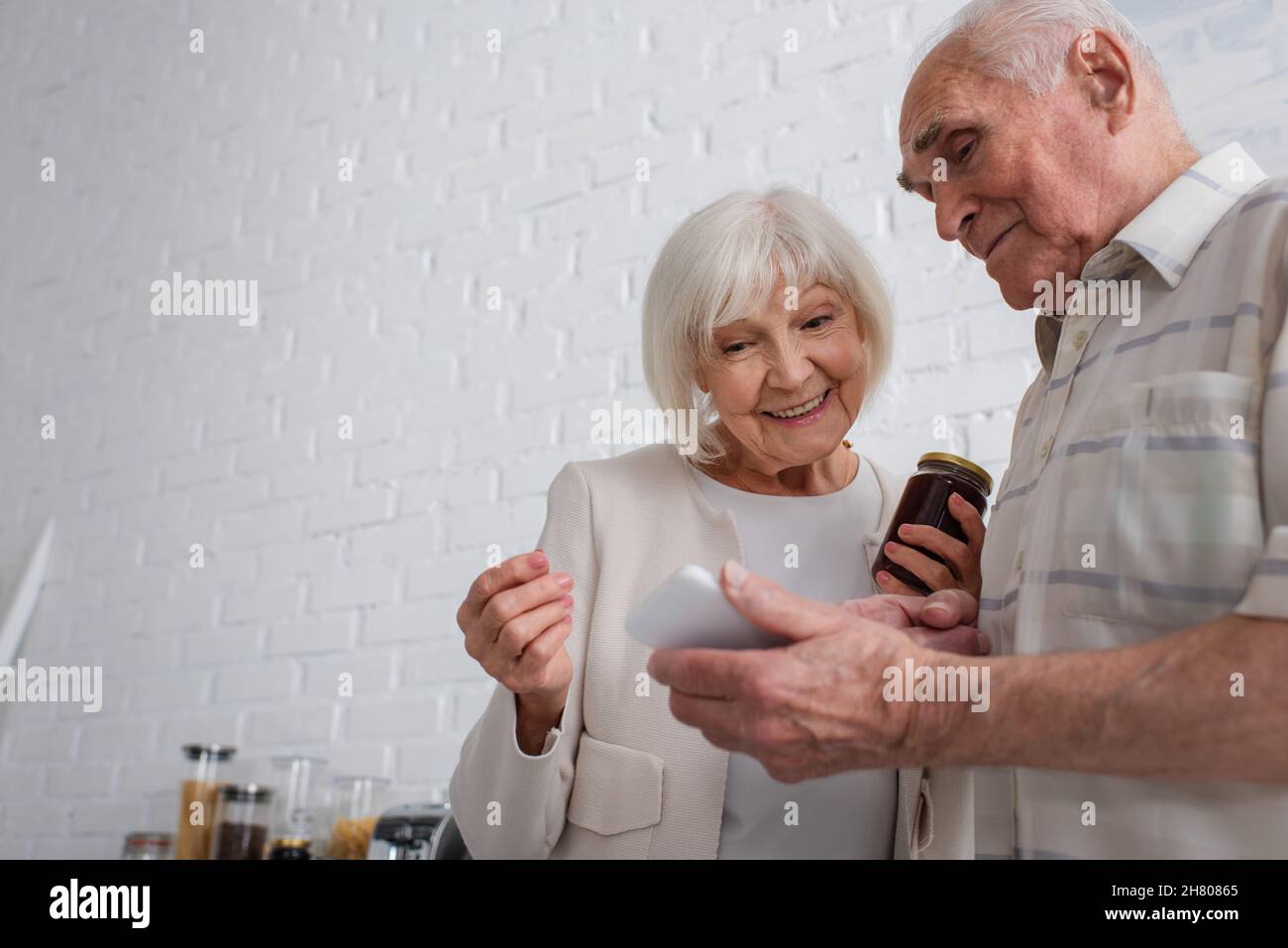 Uomo anziano che mostra smartphone a un amico sorridente con vaso di marmellata in casa di cura Foto Stock
