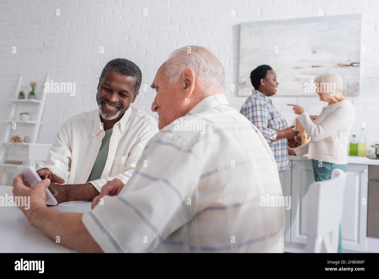 Gli uomini anziani allegri usando lo smartphone vicino a blurred gli amici multietnici nella casa di cura Foto Stock