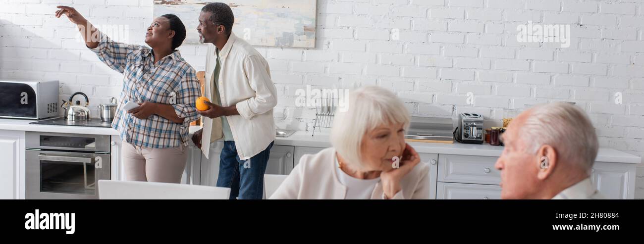Donna afroamericana senior con smartphone puntando con dito vicino amico con frutta in casa di cura, banner Foto Stock