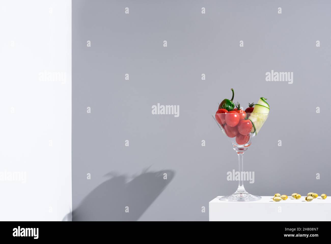 Pomodori ciliegini con cetriolo e pepe verde in bicchiere da cocktail su supporto su sfondo bianco in studio con luce solare Foto Stock