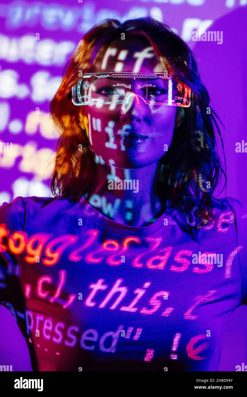 Basso angolo di giovane donna in vestito casual e occhiali a LED in piedi in luce al neon e guardando la fotocamera con codice HTML Foto Stock