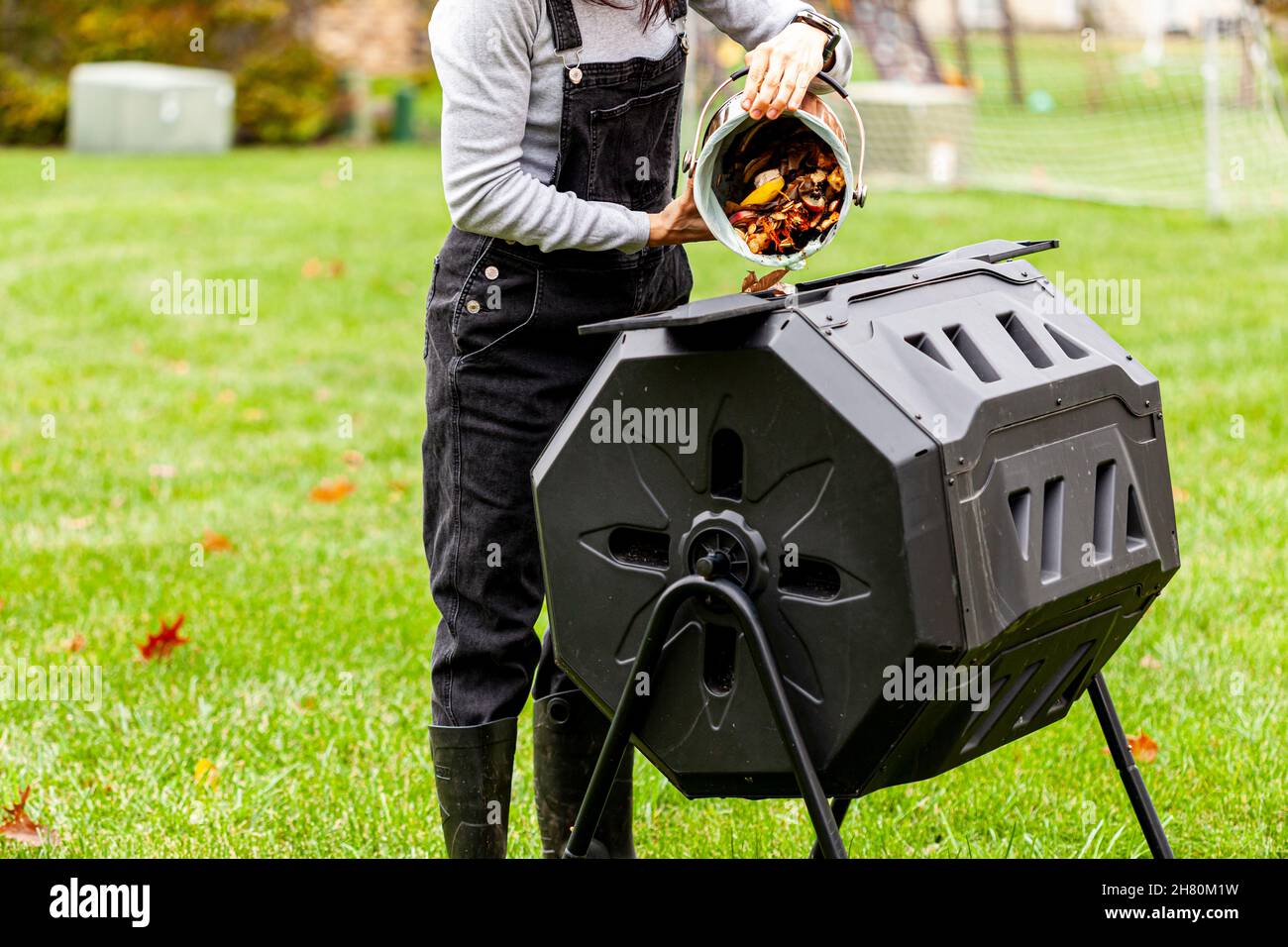 Una donna sta scaricando un piccolo bidone di scarti di cucina in un composter tumbling esterno nel giardino del cortile. Queste unità in plastica con gambe in metallo possono ruotare a. Foto Stock