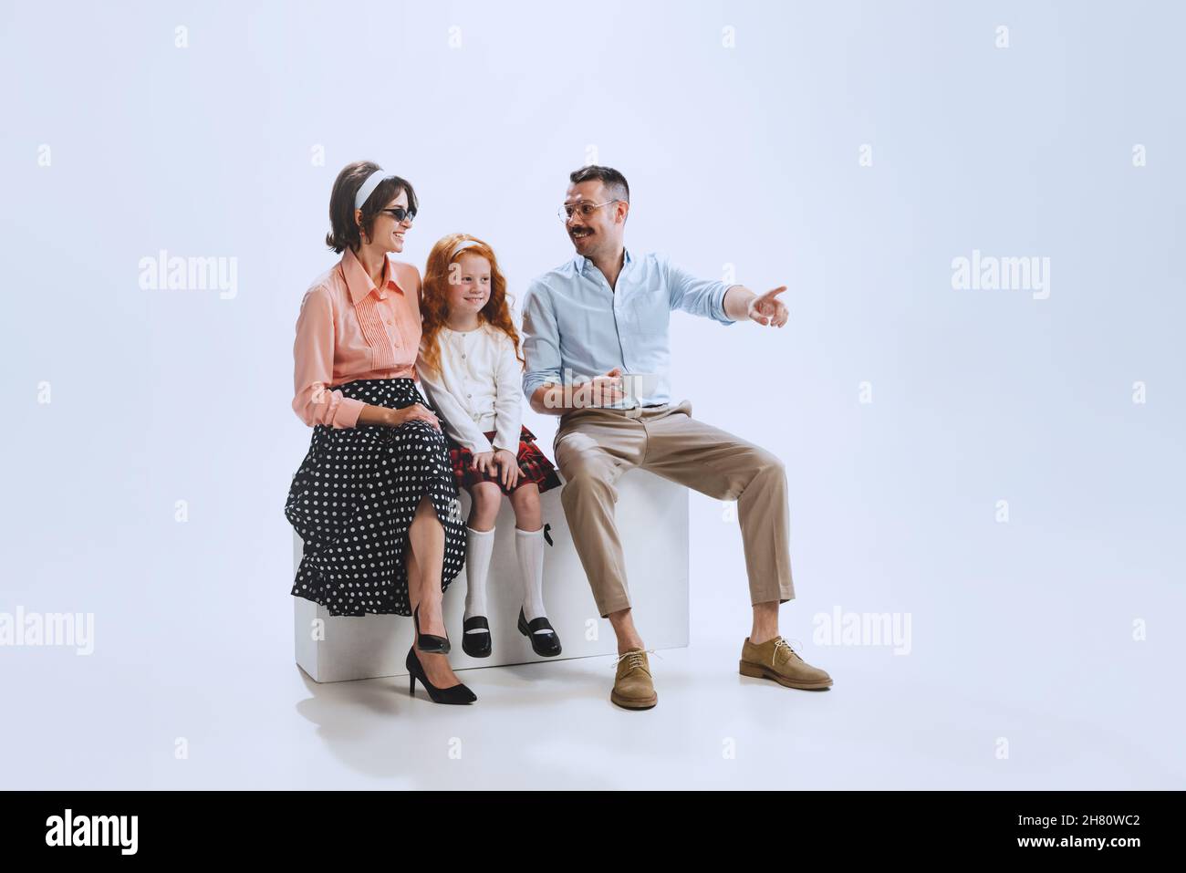 Ritratto di giovane famiglia felice, madre , padre e figlia in stile retrò abiti, moda degli anni '70, sessanta anni seduti insieme isolato su bianco Foto Stock