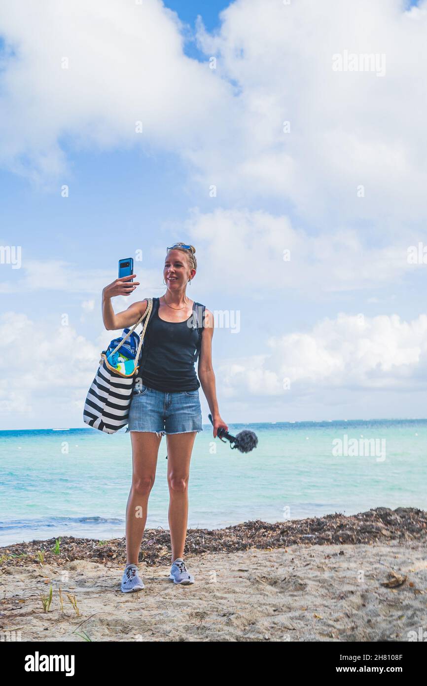 Donna prende selfie sulla spiaggia con smartphone contro le acque e le nuvole teali Foto Stock