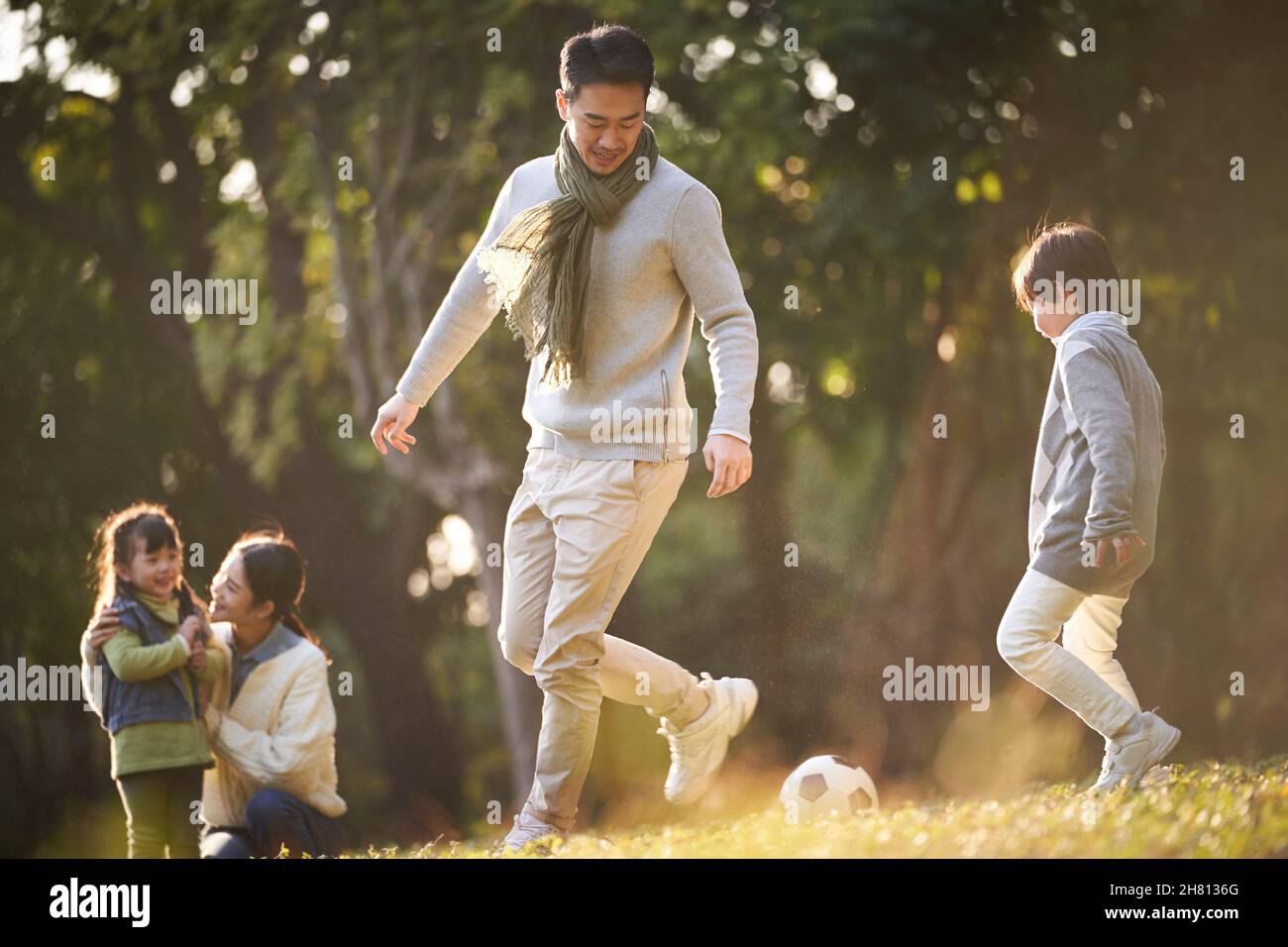 padre e figlio asiatici che giocano a calcio all'aperto nel parco, mentre madre e figlia guardano da dietro Foto Stock