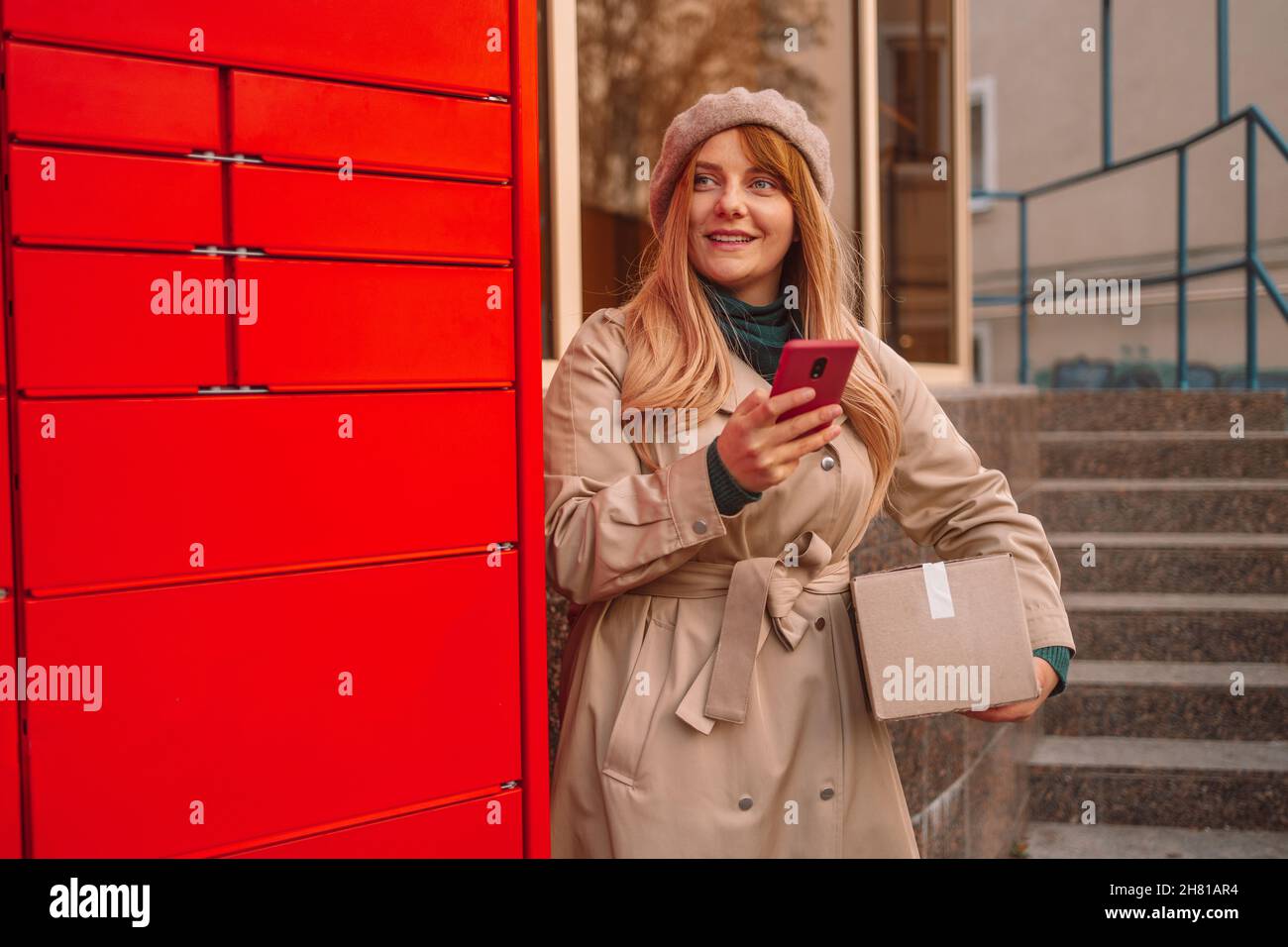 Giovane donna caucasica che tiene lo smartphone utilizzando un terminale automatico self-service o un armadietto per depositare il pacco per lo stoccaggio Foto Stock