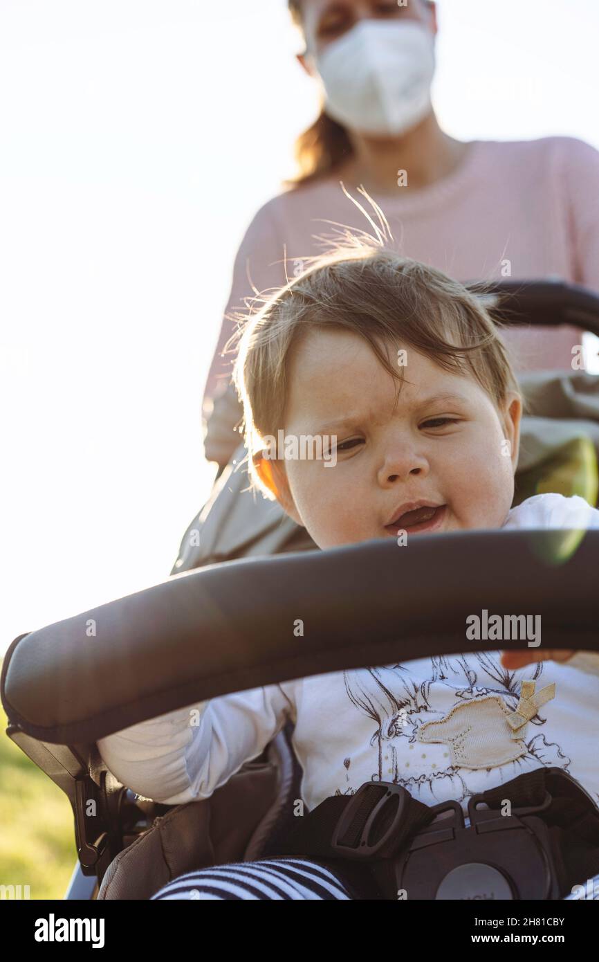 Bambina che si gode una passeggiata in un parco seduto nel passeggino moderno durante l'epidemia di coronavirus - bambino dolce in buggy - Madre con viso protettivo ma Foto Stock