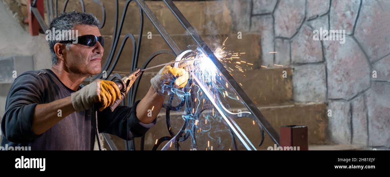 Uomo che indossa occhiali protettivi salda metallo con saldatrice in casa privata. Le scintille provenienti dalla saldatura volano sui lati. Banner. Foto Stock