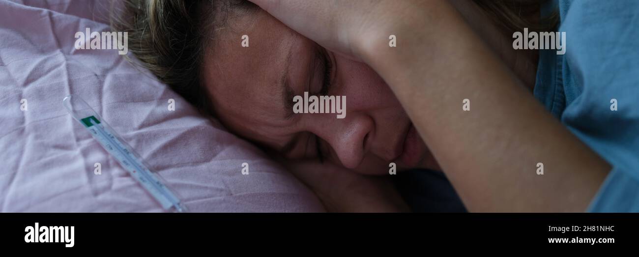Donna sdraiata a letto e che tiene la testa dolente con le mani vicino al termometro a mercurio Foto Stock