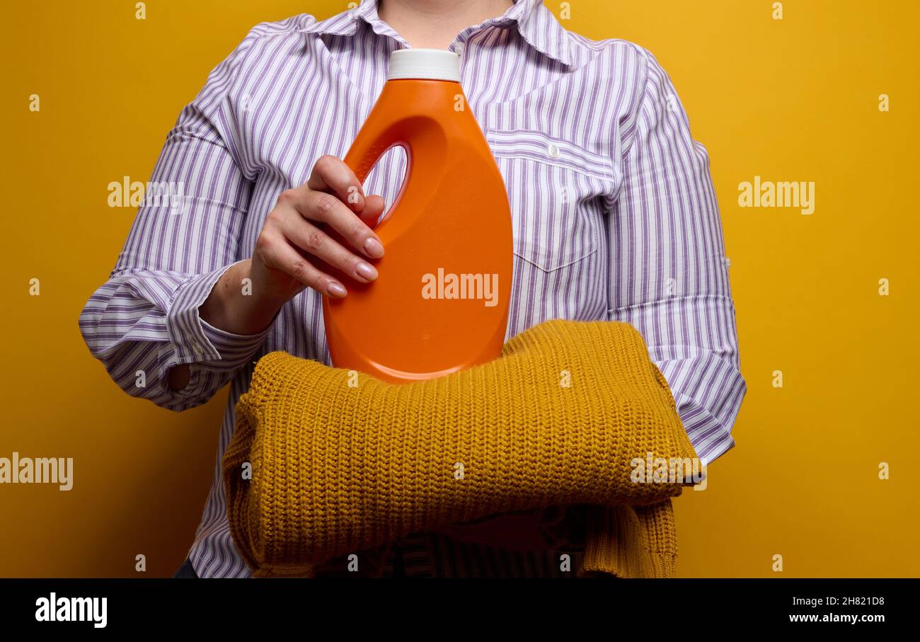 una donna in una camicia a righe bianche tiene una bottiglia di plastica arancione con gel liquido per lavaggio e un maglione a maglia. La lavanderia e le faccende domestiche, era pulita Foto Stock