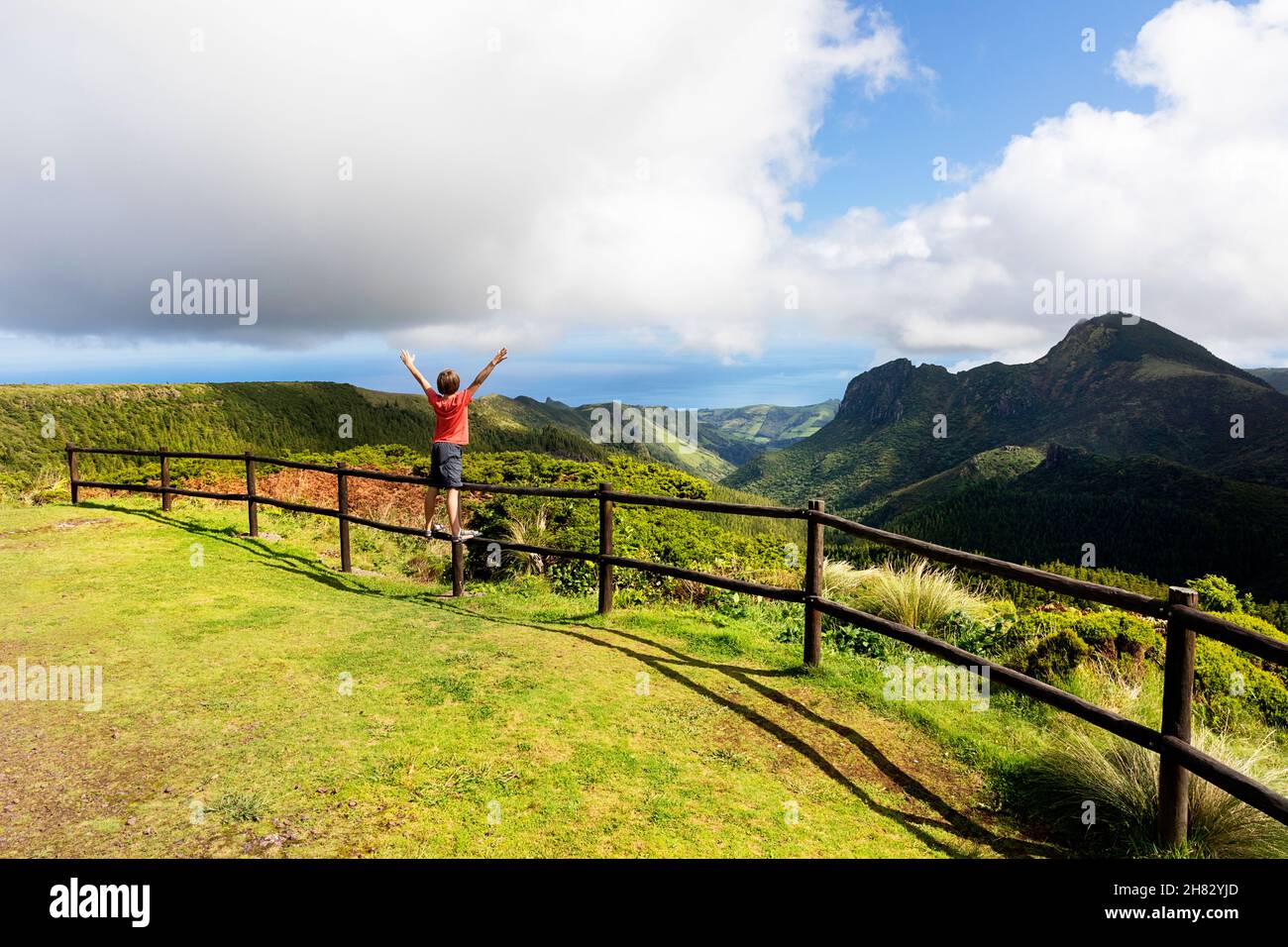 Ragazzo in t shirt rossa standin su recinzione di legno alzando le mani in aria al punto di vista Miradouro do vale da Fazenda a Flores, Azzorre, Portogallo Foto Stock