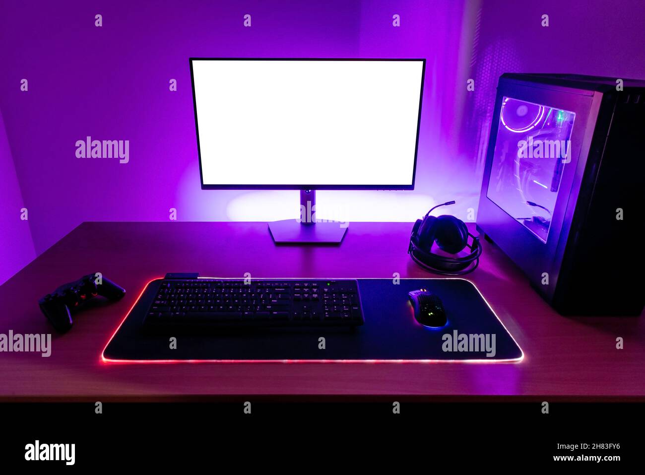Scrivania con configurazione per giochi. Display con schermo isolato per mockup. PC da gioco, cuffie, tastiera, mouse e joypad sulla scrivania. Luce a LED viola sulla parete Foto Stock