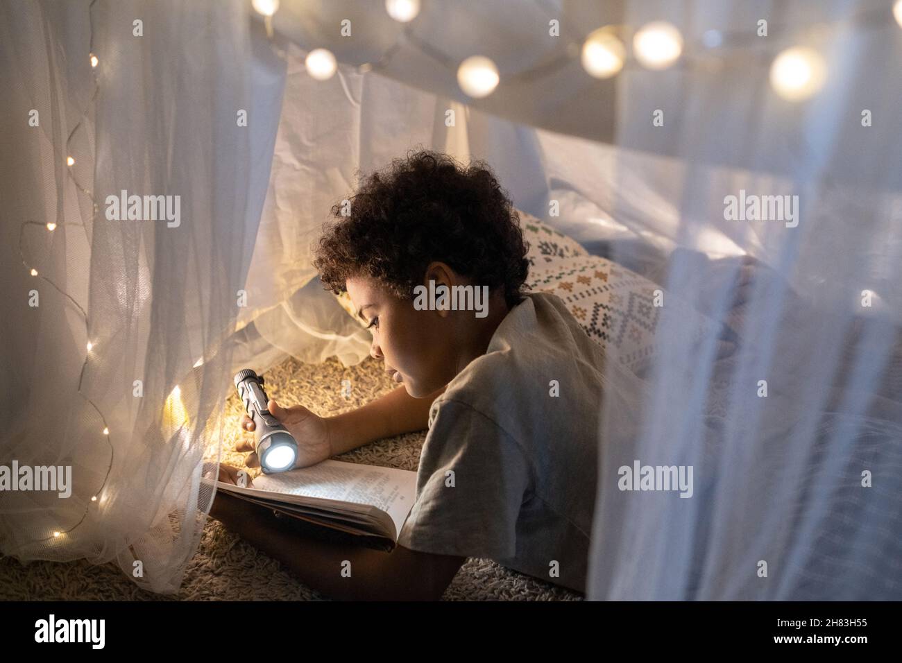 Curioso ragazzo nero con capelli ricci che giacciono all'interno della tenda da gioco decorata con luci di Natale e libro di lettura con torcia Foto Stock