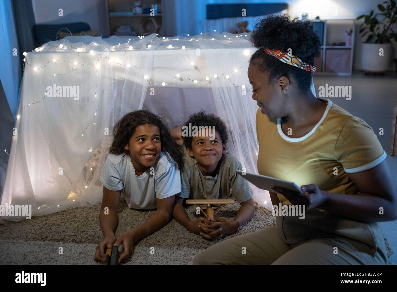 Donna nera sorridente usando il tablet mentre legge il libro in linea ai bambini all'interno della tenda di gioco prima del letto Foto Stock
