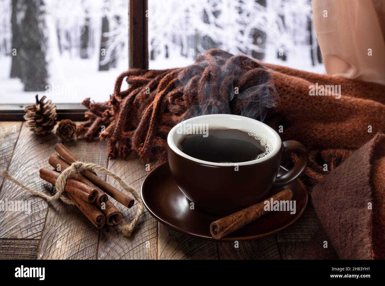 Una tazza di caffè fumante su un tavolo di legno vicino a una finestra con sfondo invernale Foto Stock