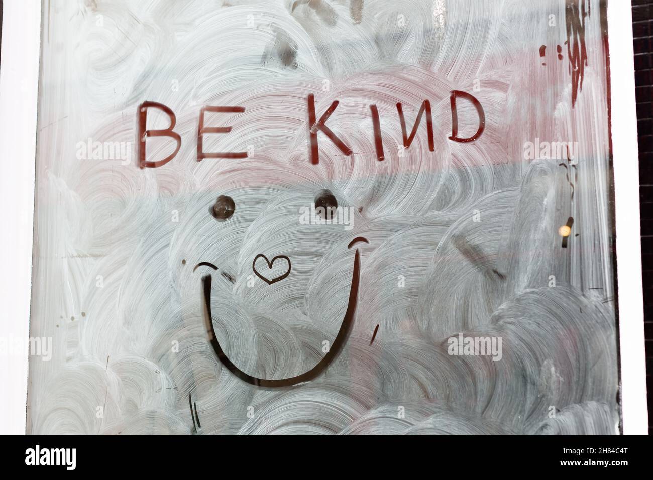'Siate gentili' con un viso sorridente e cuore d'amore disegnato in bianco lavaggio su una vetrata vuota per diffondere un po' d'amore e positività durante la pandemia Foto Stock