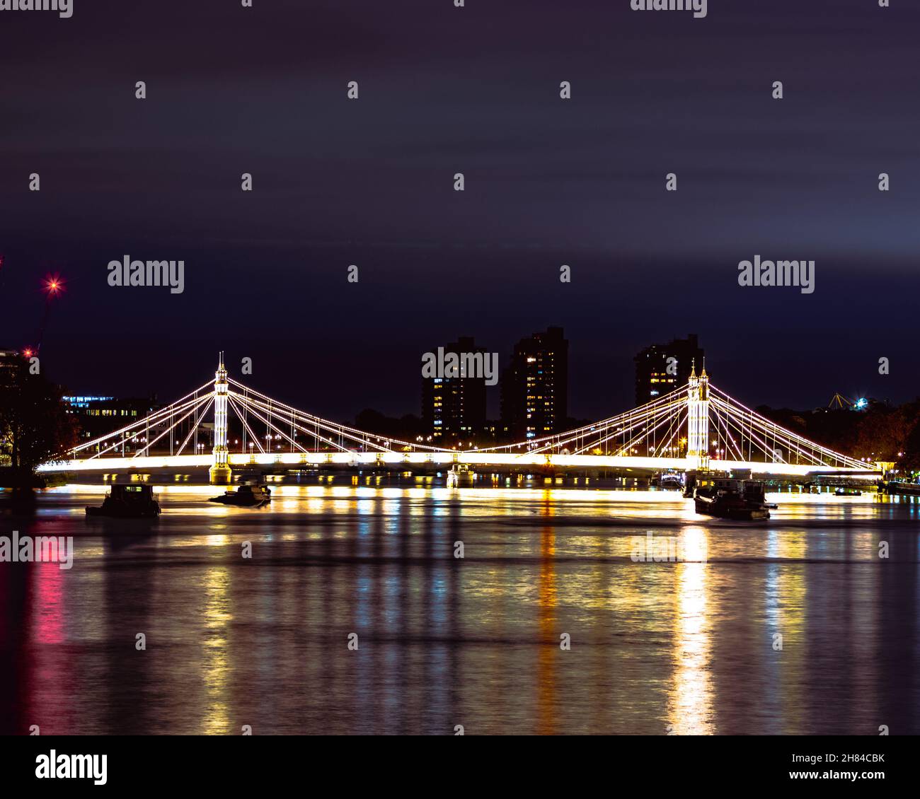 Albert Bridge illuminato di notte con grattacieli sullo sfondo e il tamigi in primo piano. Girato dal ponte di Chelsea. Foto Stock