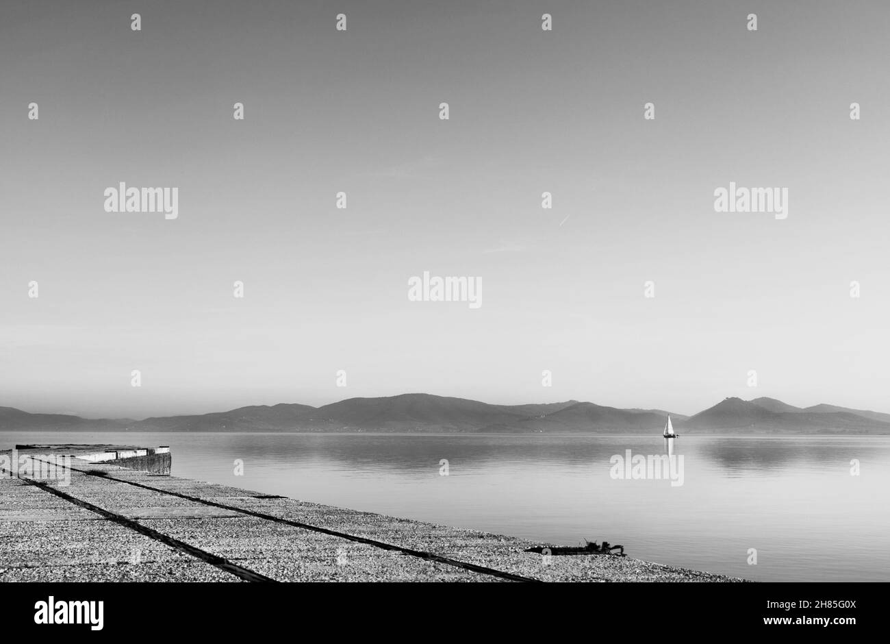 Una barca a vela nelle calme acque del Lago Trasimeno , Italia , in primo piano costa con piccole città , fotografia in bianco e nero Foto Stock