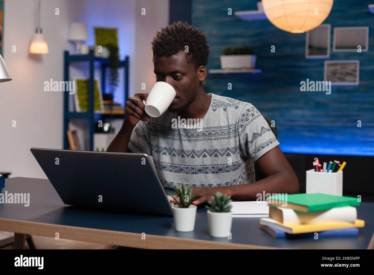 Dipendente afroamericano che tiene una tazza di caffè digitando idee di marketing sul computer che lavora in remoto al progetto online in salotto. Freelance seduto alla scrivania che analizza il grafico finanziario Foto Stock