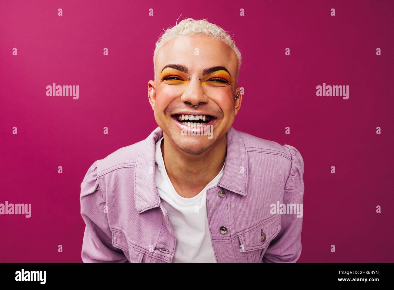 Un queer uomo sicuro che sorride alla fotocamera mentre si alza in piedi da solo in uno studio. Giovane uomo non conforme con occhi colorati e gioielli di fronte Foto Stock