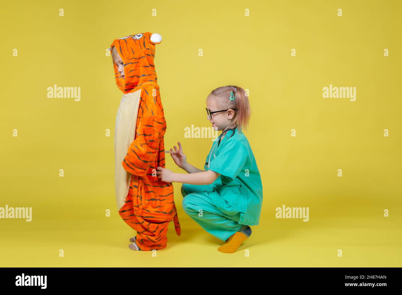 I bambini caucasici piccoli giocano il medico, una ragazza vestita come medico dà un'iniezione ad un ragazzo vestito come tigre Foto Stock