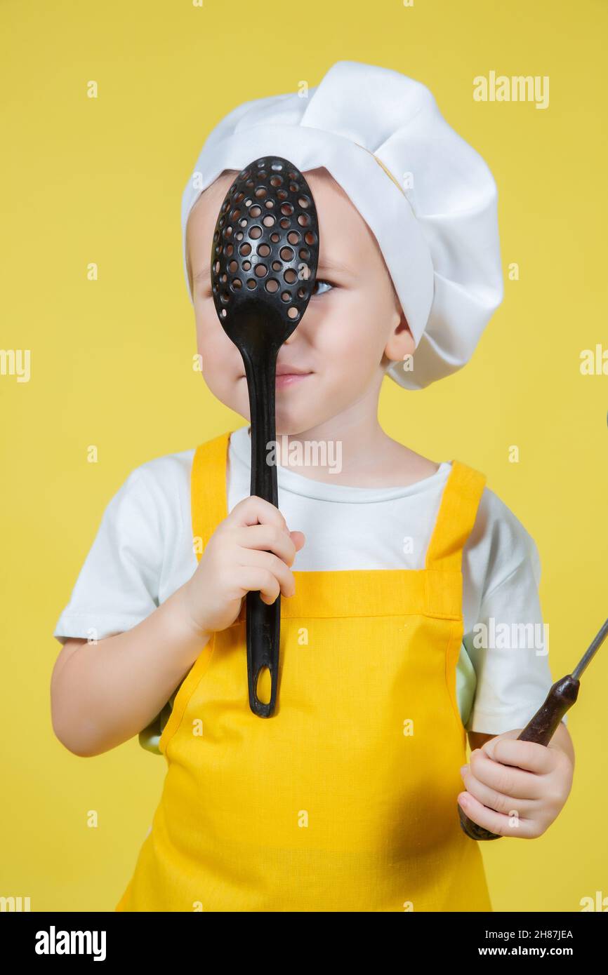 Piccolo ragazzo caucasico che gioca chef, ragazzo in grembiule e cappello dello chef che si posa su sfondo giallo Foto Stock