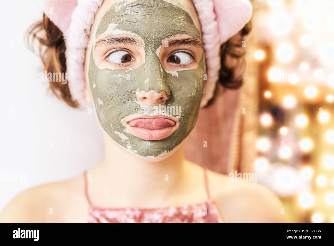 ritratto di una ragazza adolescente con capelli marroni con una maschera cosmetica di argilla grigia sul suo viso con una simpatica grimace. Umore allegro, faccia i volti, occhi inclinati. Foto Stock