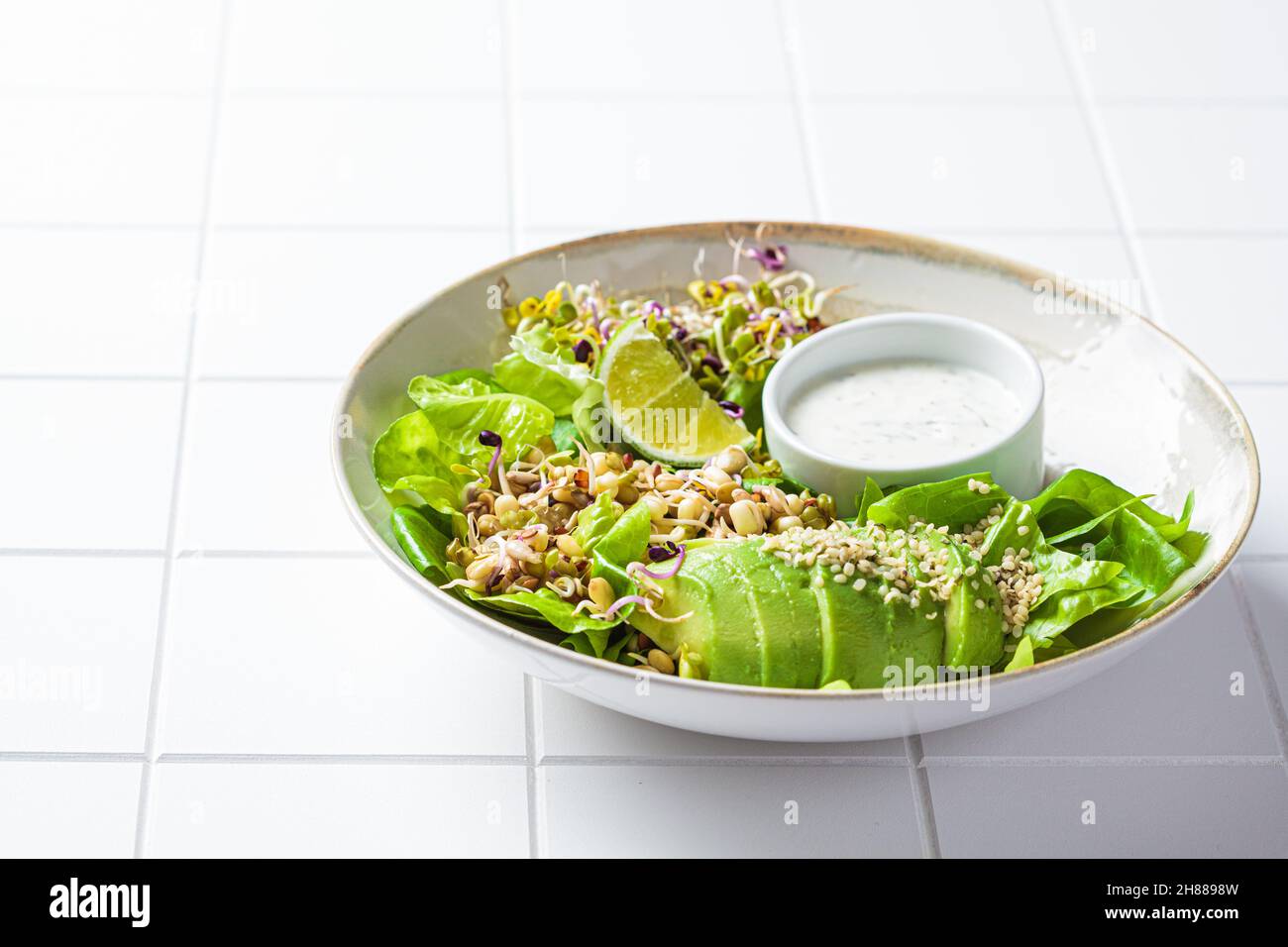 Insalata di disintossicazione verde con avocado, germogli, semi di canapa e condimento yogurt, sfondo piastrellato bianco, spazio copia. Foto Stock Insalata di disintossicazione verde con avocado, germogli, semi di canapa e condimento yogurt, sfondo piastrellato bianco, spazio copia. Foto Stock