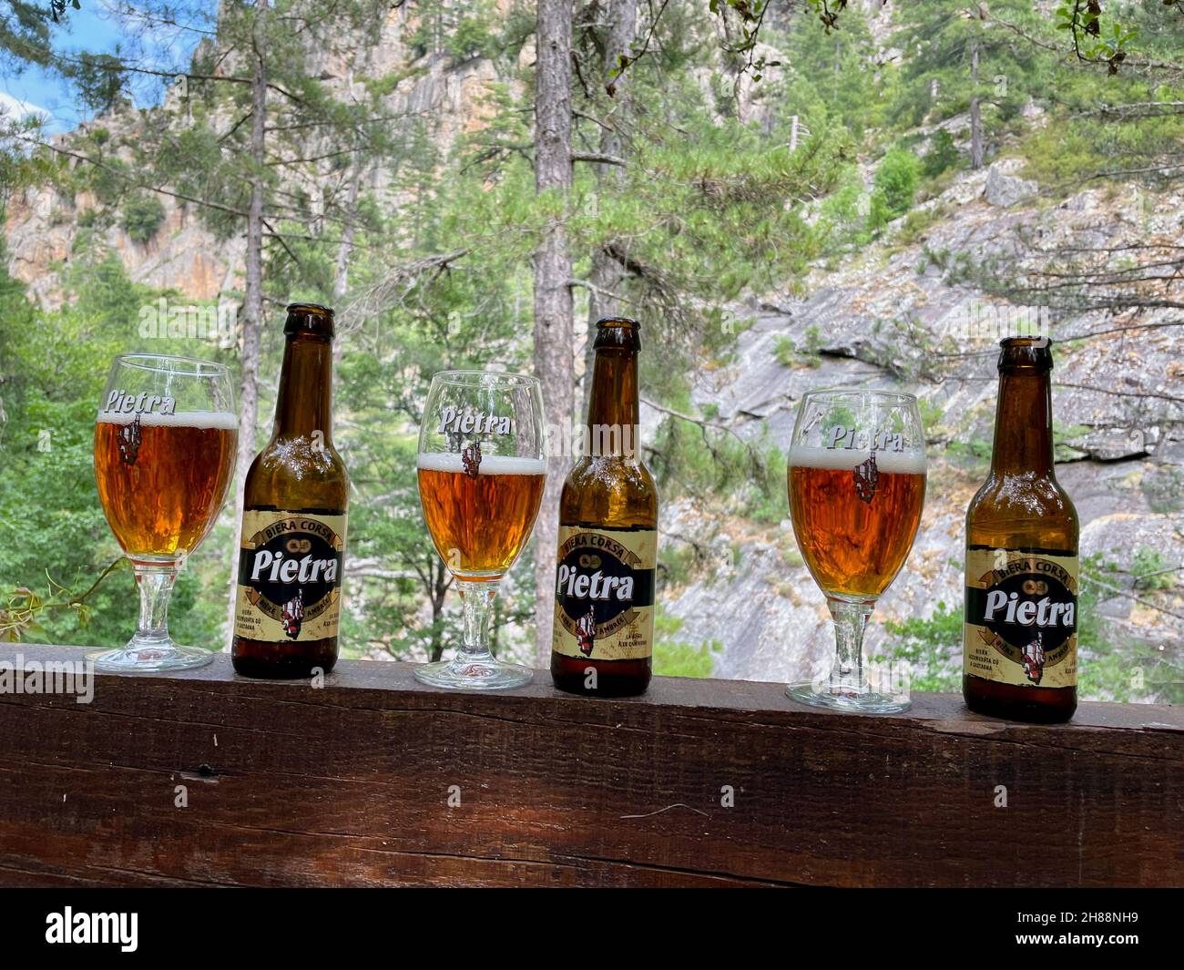 Valle Restonica, Corsica, 21.07.2021. Tre bicchieri da birra pietra e bottiglie in piedi su ringhiera di legno con pini e rocce sullo sfondo. Foto Stock