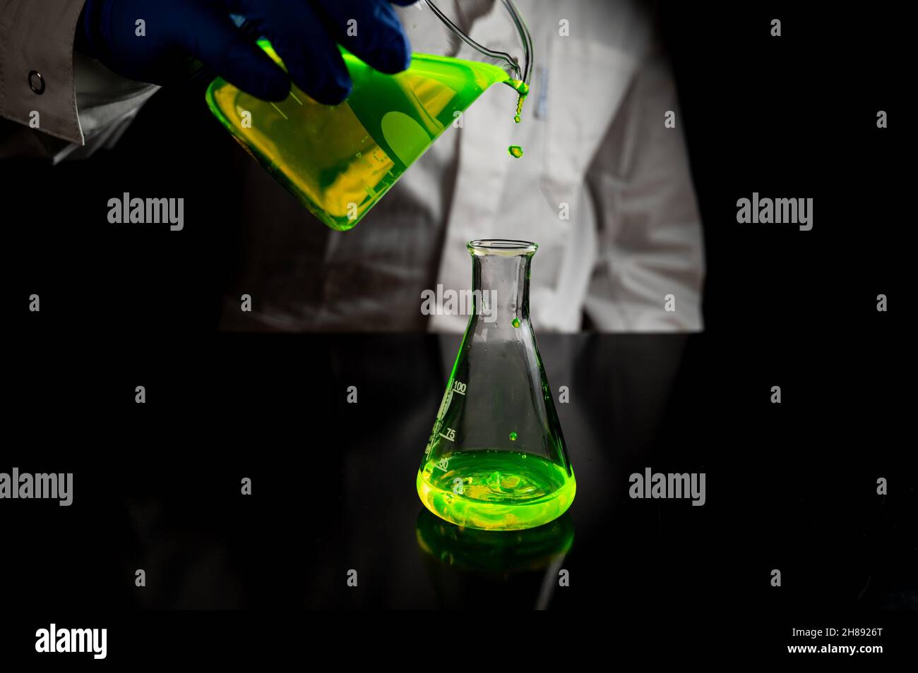 Uno scienziato di ricerca che sperimenta con gocce fluorescenti verdi in un pallone conico di vetro in laboratorio biomedico scuro per medicina di cura della salute d Foto Stock