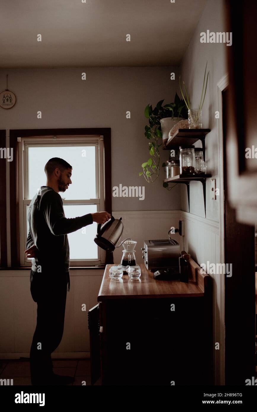 Il giovane uomo millenario di ippster misto-corsa eroga caffè a casa utilizzando un pour-over in cucina, piante, finestra, moody editing Foto Stock