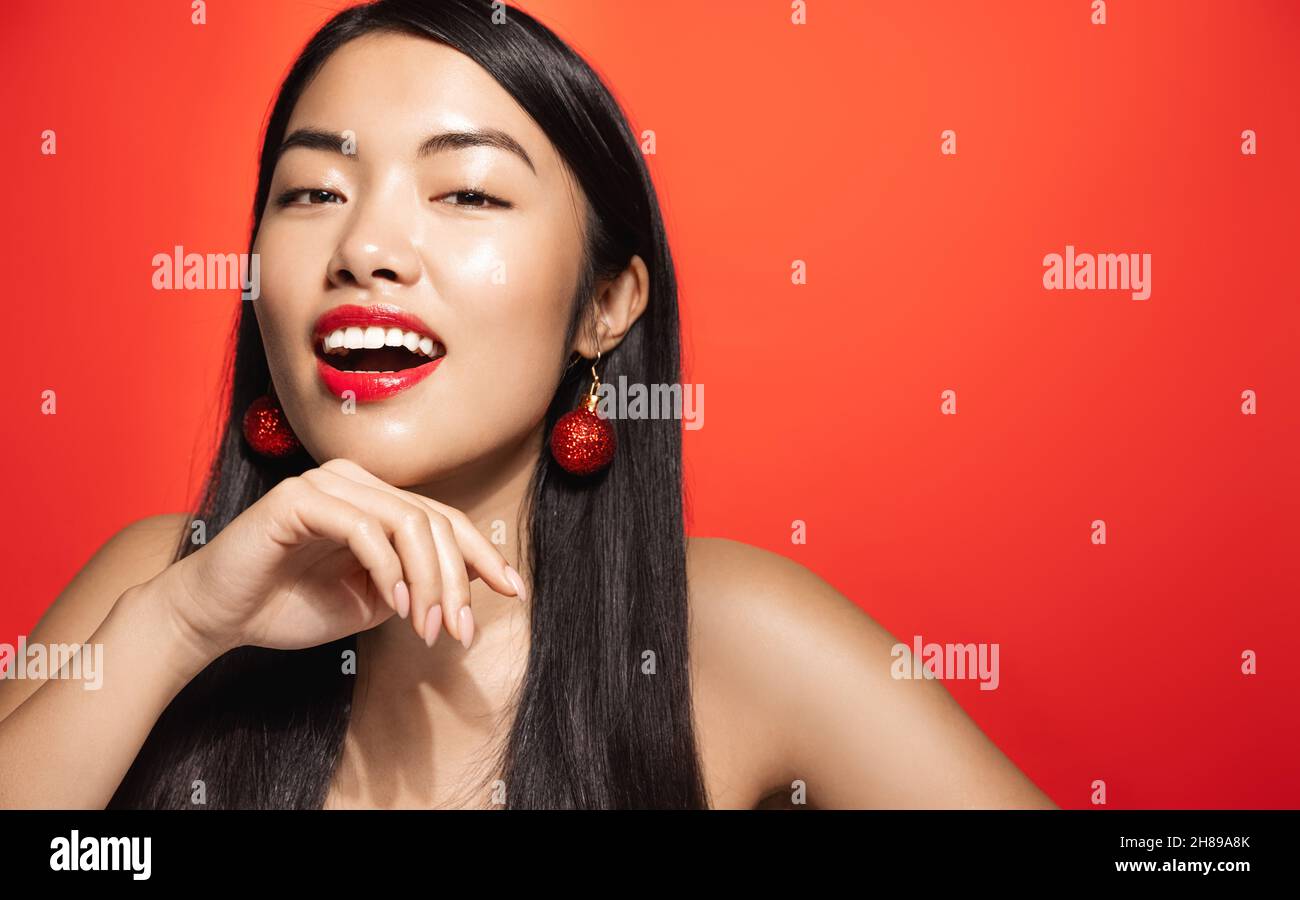 Ritratto di donna asiatica sexy con arredamento orecchini di natale, sorridente e sguardi sensuale, sorridente alla macchina fotografica, mostrando trucco e pelle chiara, rosso Foto Stock