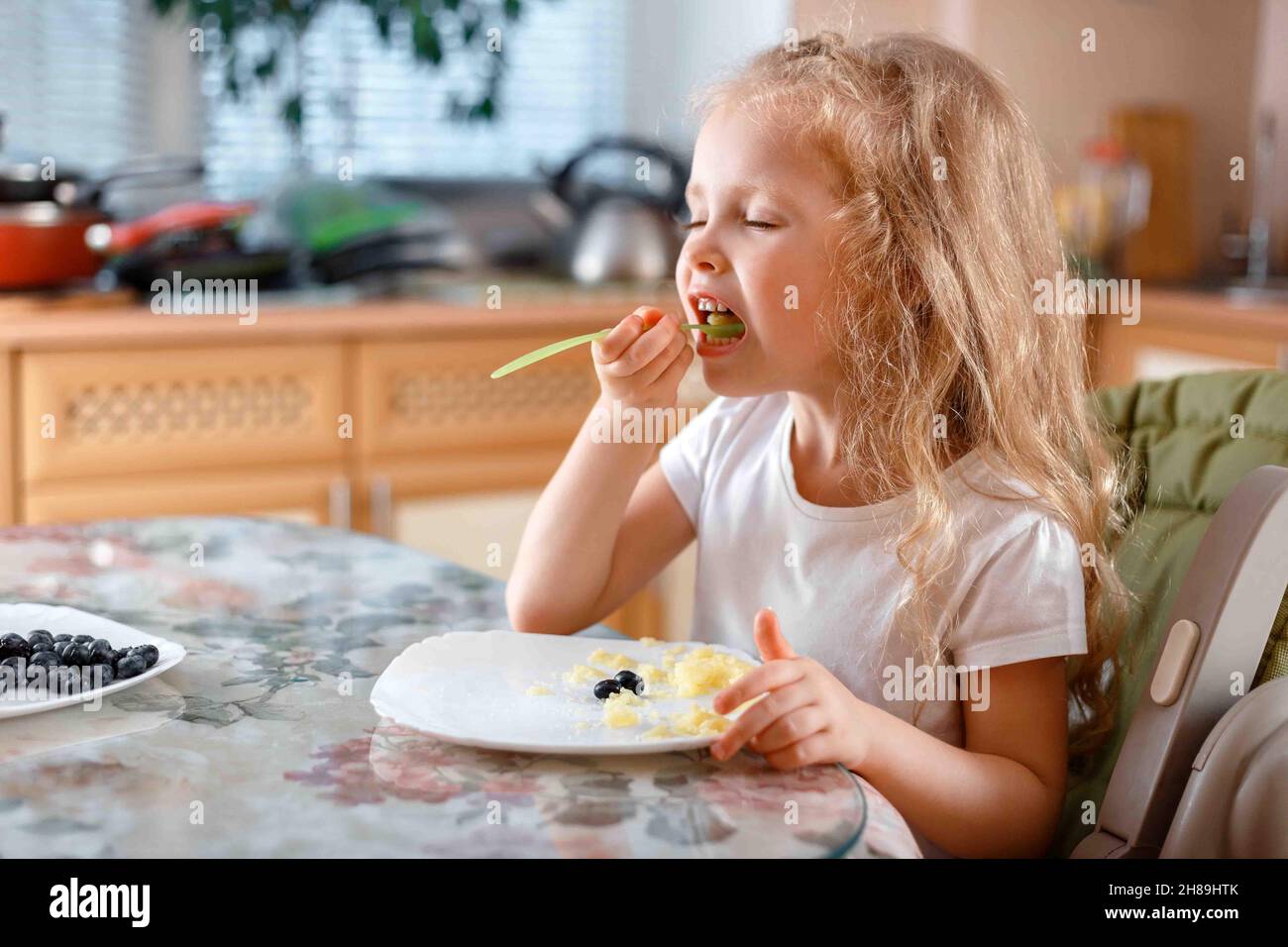 Bambina bambino che mangia porridge con bacche in cucina al tavolo da cucchiaio di plastica seduto sulla sedia per alimenti per bambini. Pranzo di allegro capretto caucasico biondo Foto Stock