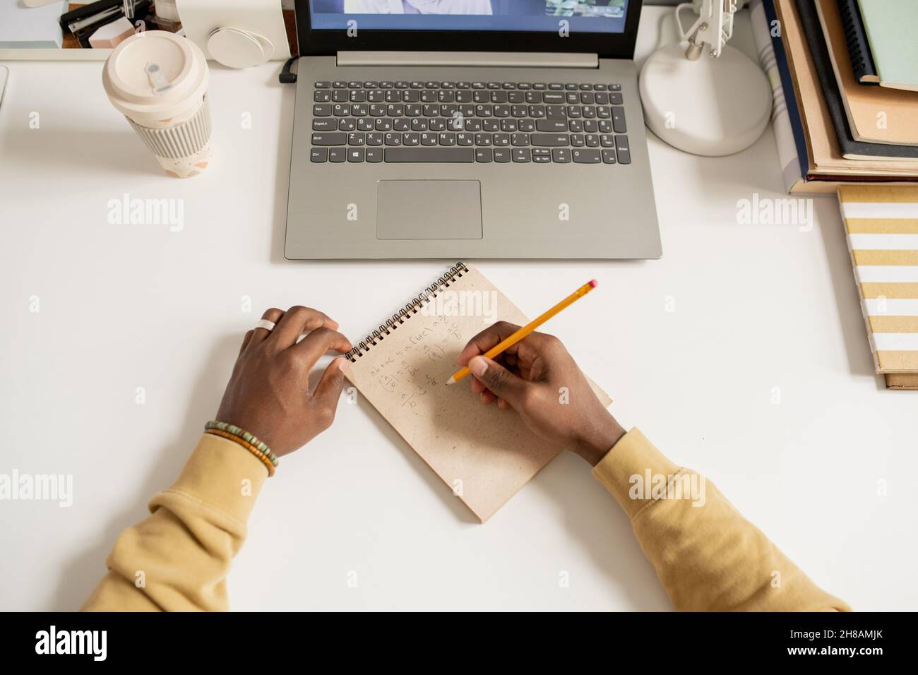Mani di giovane africano scrivere formula algebrico a pagina di blocco note mentre si siede dalla scrivania di fronte al computer portatile Foto Stock
