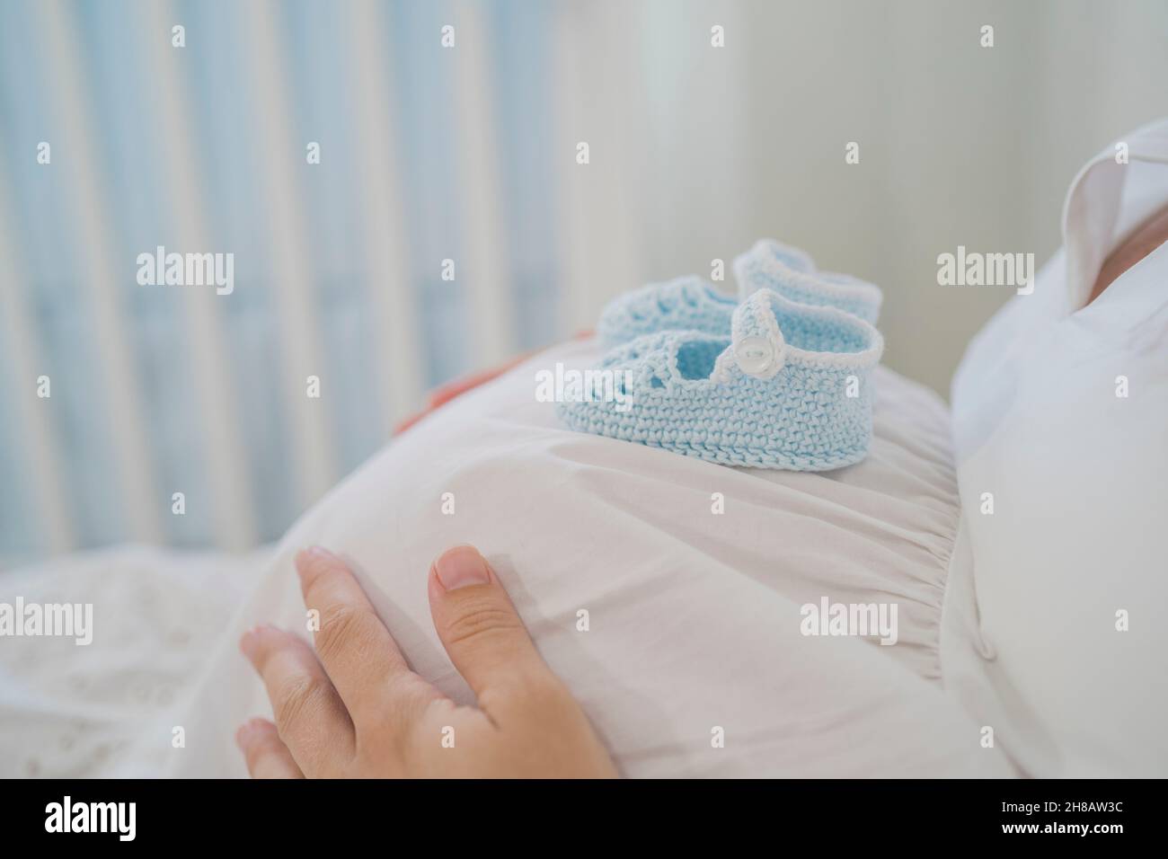 Crop donna incinta senza volto con scarpe per bambini sul pancino che giace sul letto Foto Stock